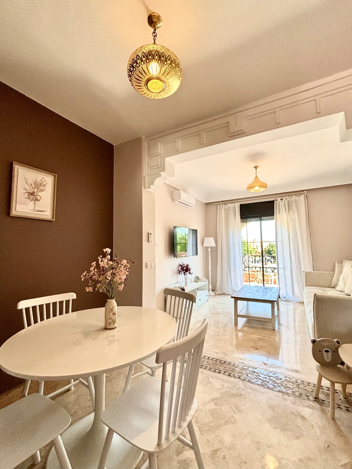 A dining area is presented with a round white table and four chairs. A soft floral arrangement is positioned in the center. Light streams through large windows adorned with sheer curtains, casting a gentle glow on the beige walls and marble flooring.