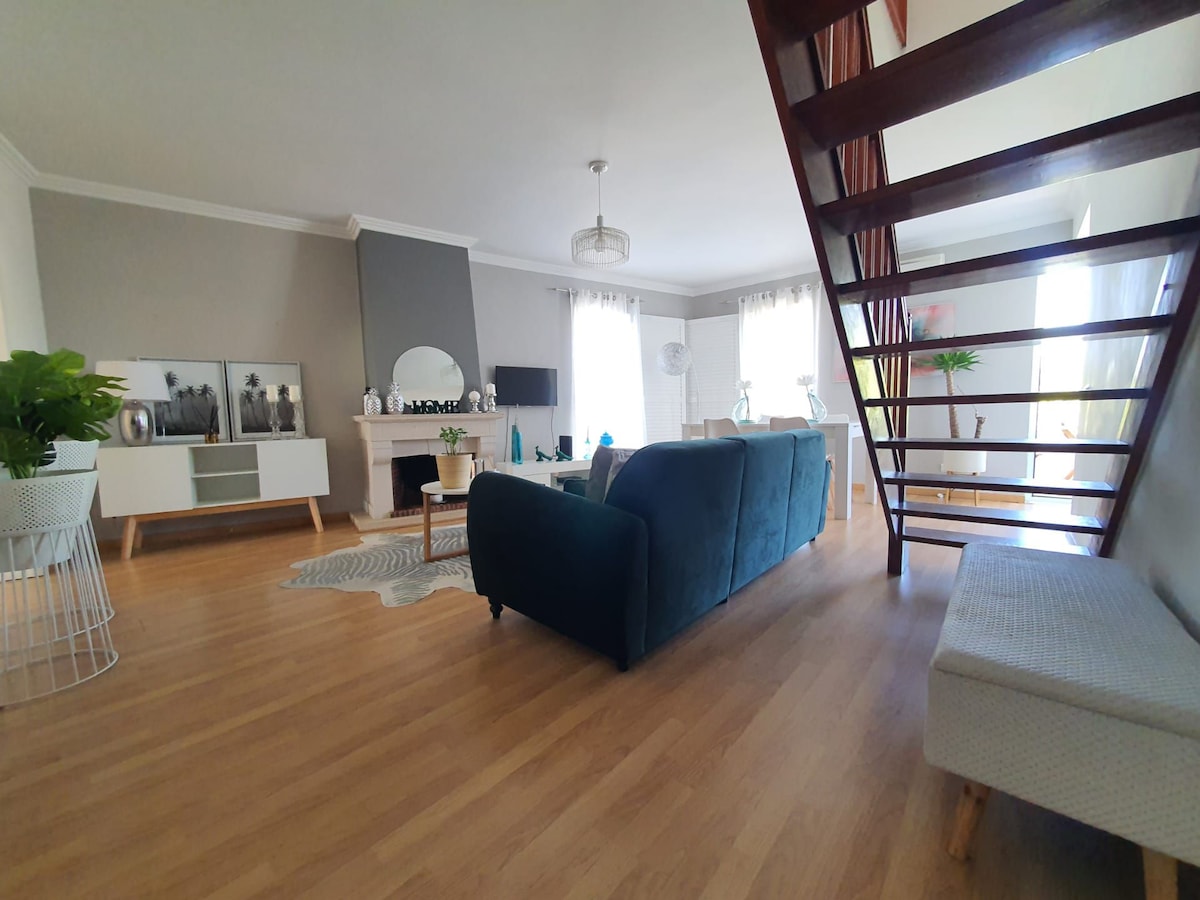 A spacious living area features a comfortable blue sofa, complemented by a light wooden floor. A decorative rug lies underneath a coffee table, and a bright staircase leads to the upper level. Natural light floods the room through large windows, enhancing the welcoming atmosphere.