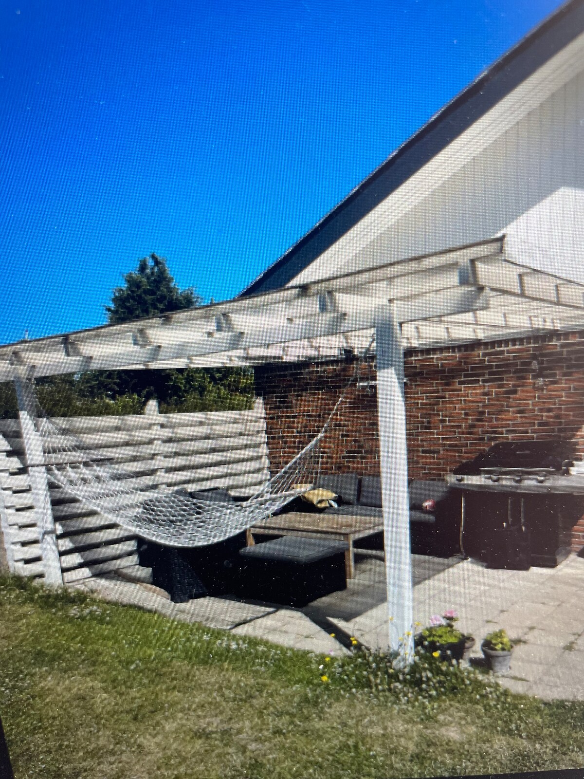 A shaded outdoor area features a comfortable seating arrangement with dark cushions, a hammock, and a barbecue grill. Natural grass and small flowers surround the stone-paved patio, while a wooden fence provides privacy.