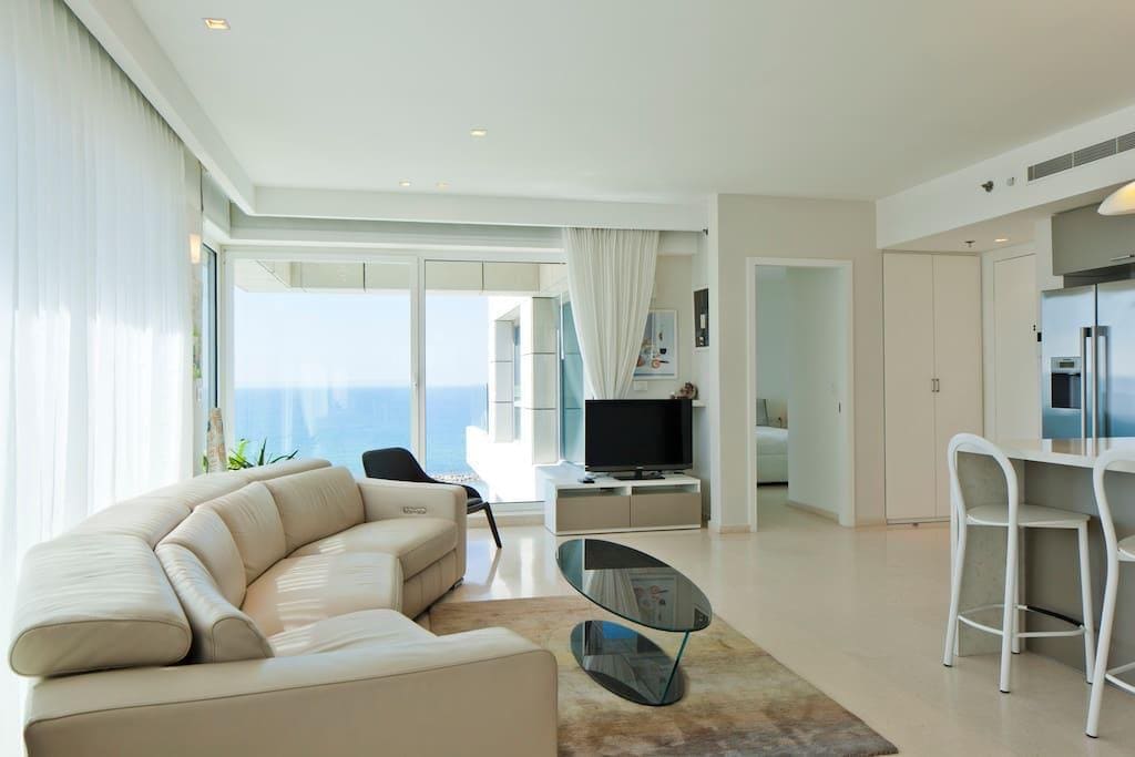 A modern living area is highlighted by a curved sectional sofa and a glass coffee table. Large windows offer expansive views of the sea, while natural light illuminates the space. A television is positioned opposite the seating area, enhancing the sense of comfort.