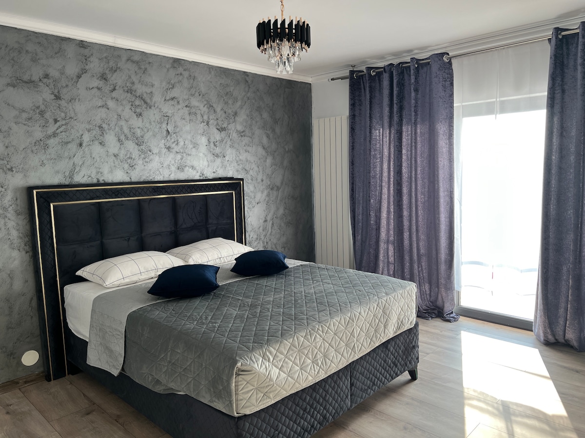 A contemporary bedroom features a spacious bed dressed in light linens with navy accent pillows. Gray walls provide a modern backdrop, while long, dark curtains frame a balcony door that allows natural light to fill the room.