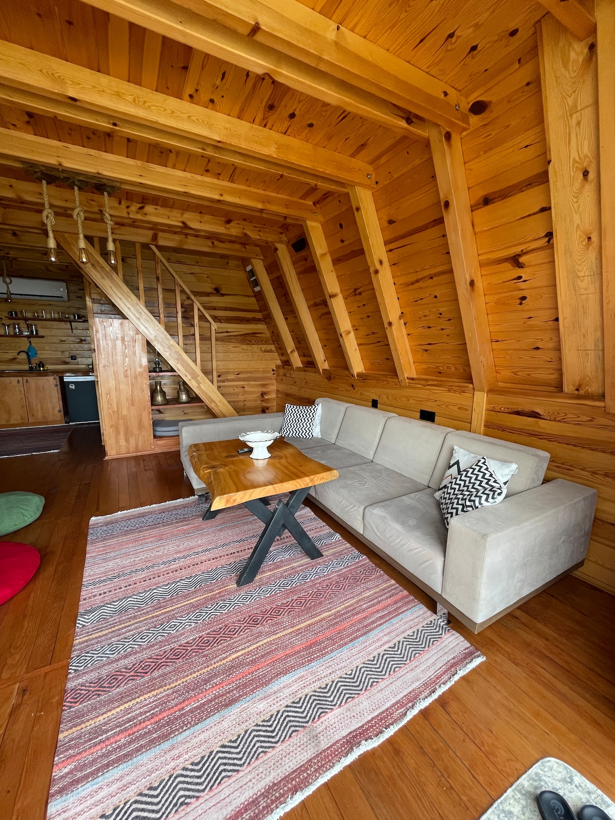 A comfortable seating area features a beige sofa paired with a wooden coffee table, set on a patterned rug. The warm wooden walls and exposed beams contribute to a cozy atmosphere. A staircase is visible in the background, leading to the upper level.