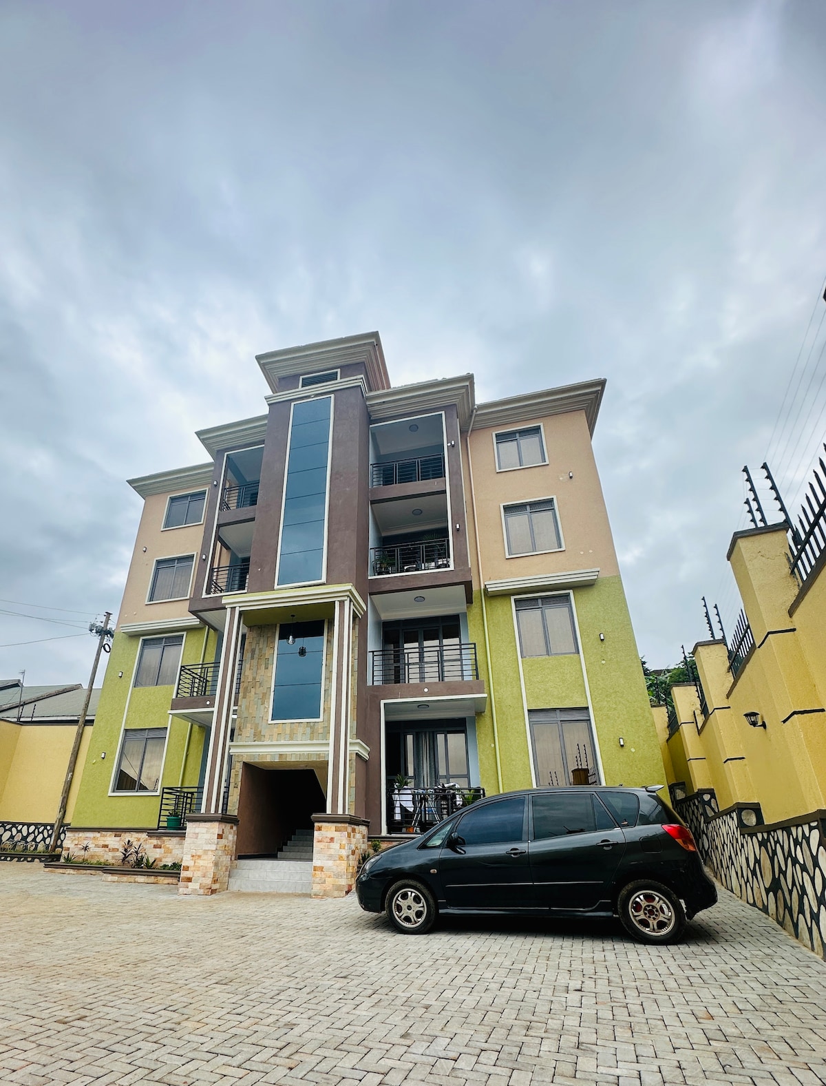 A modern four-story building stands prominently in the image, featuring a mix of green, tan, and brown exterior finishes. Large glass windows are visible on the front, providing a glimpse of the interior. A black car is parked in the driveway paved with stones.