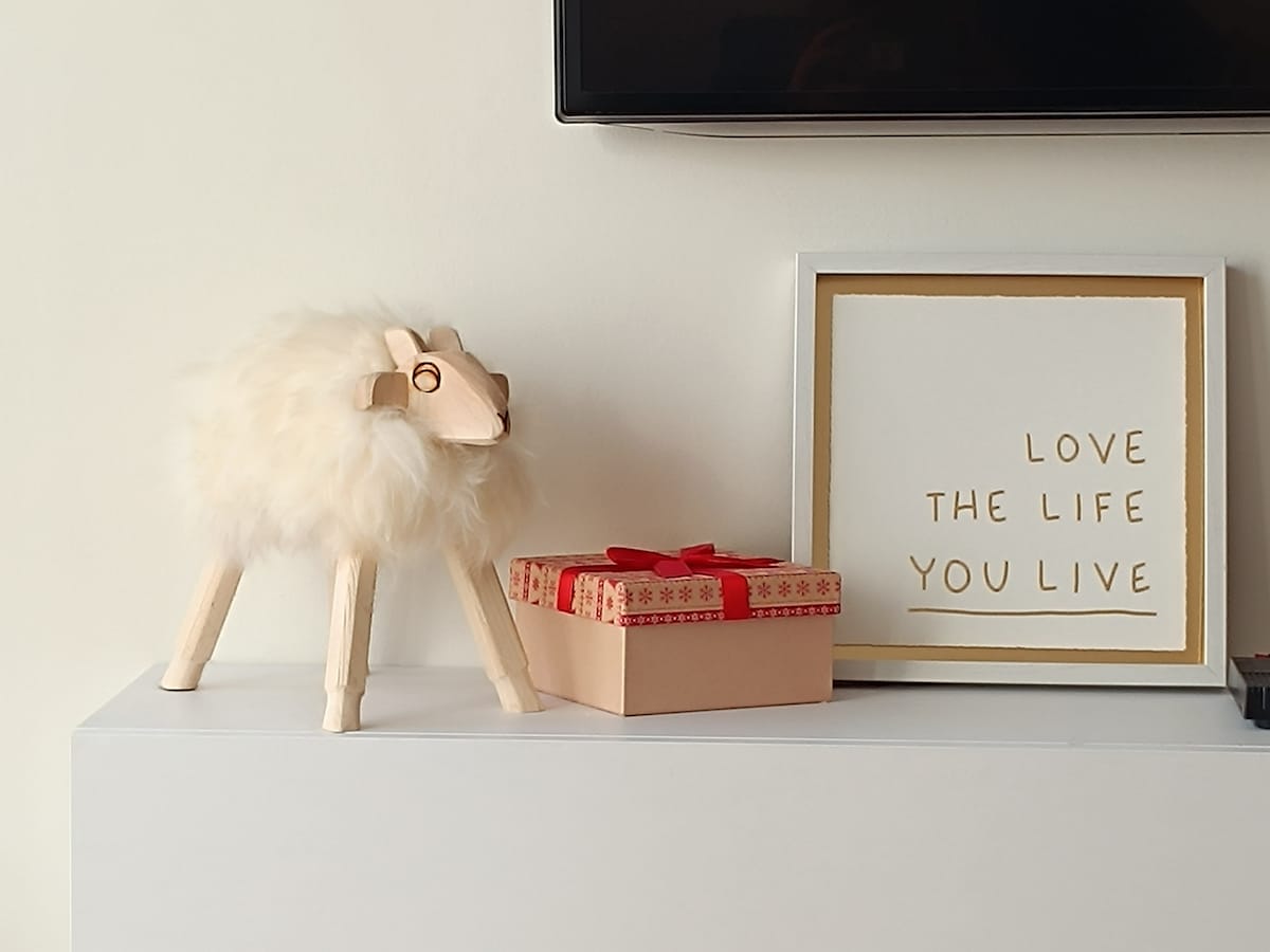A decorative arrangement is presented on a white shelf, featuring a plush sheep figurine and a gift box tied with a red ribbon. A framed artwork with the phrase "LOVE THE LIFE YOU LIVE" is displayed alongside, offering a touch of charm to the setting.