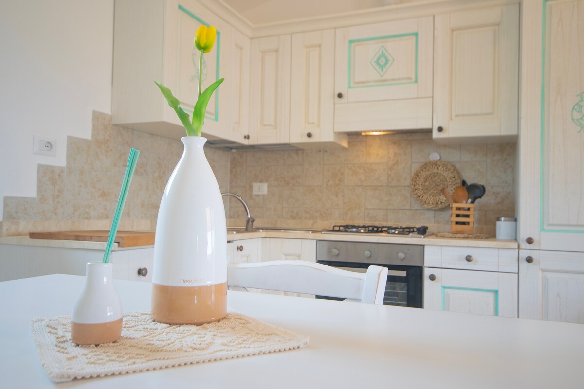 A modern kitchen area features white cabinetry with decorative accents. A white vase with a yellow tulip is placed on a textured table mat, beside a small container. Multiple kitchen appliances and utensils are arranged neatly in the background, hinting at an inviting cooking space.