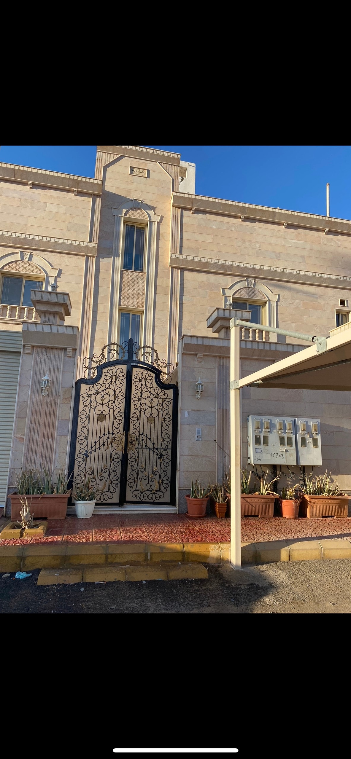 The exterior of a multistory stone building is showcased, featuring an ornate wrought iron gate at the entrance. Potted plants line the path leading to the door, and a shaded area is visible to the side, providing a welcoming entryway.