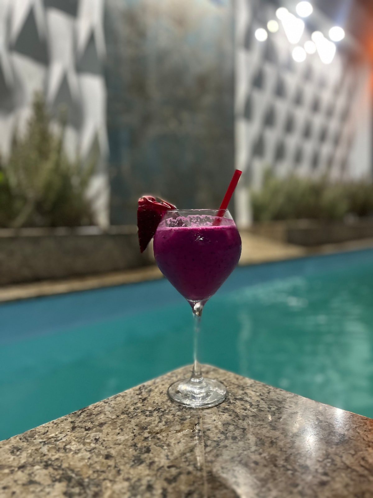 A vibrant tropical drink is presented in a stemmed glass, featuring a deep purple hue and garnished with a fresh strawberry. The drink is placed on a polished surface, with a turquoise pool visible in the background, reflecting soft lighting from above.