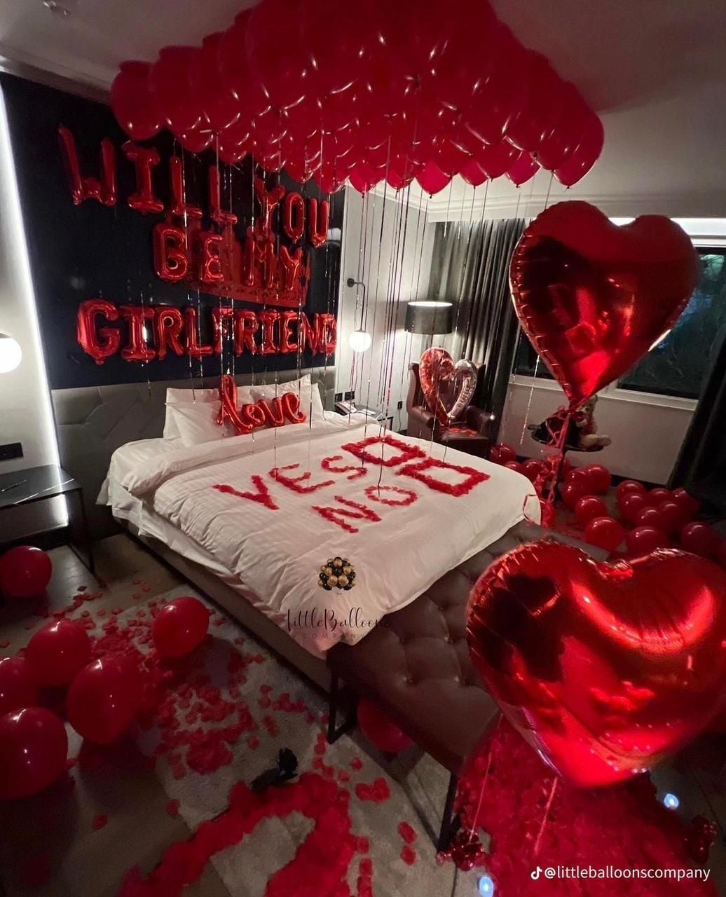 A romantic bedroom is decorated with red heart-shaped balloons and plush rose petals scattered across the floor. The spacious bed features a white duvet with a message in red letters. Romantic lighting is provided by bedside lamps, creating an intimate setting.