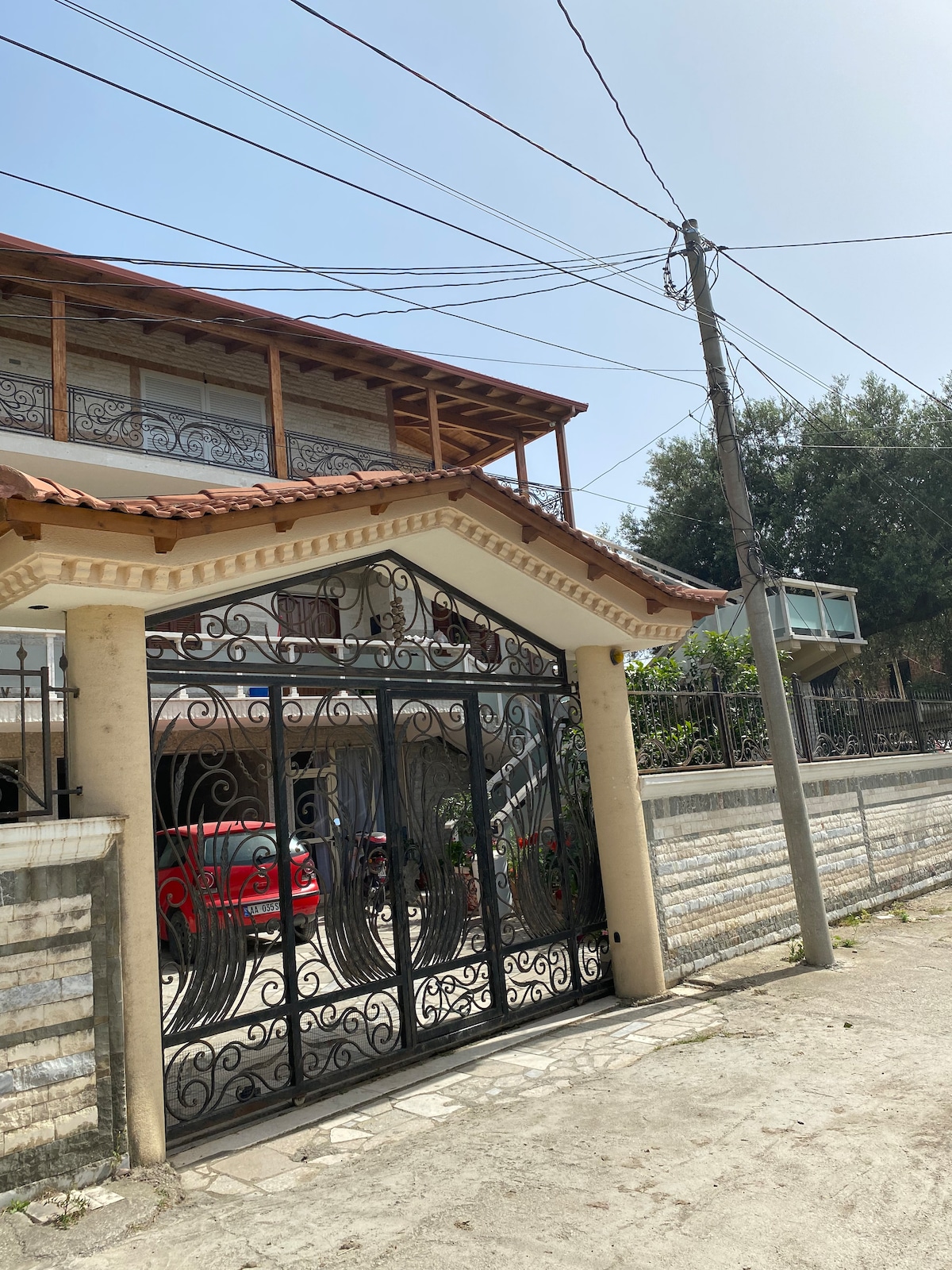 An ornate wrought iron gate stands at the entrance of a two-story house with a tiled roof. The exterior features stone walls, and a red car is parked in the driveway. Power lines are visible above against a blue sky.