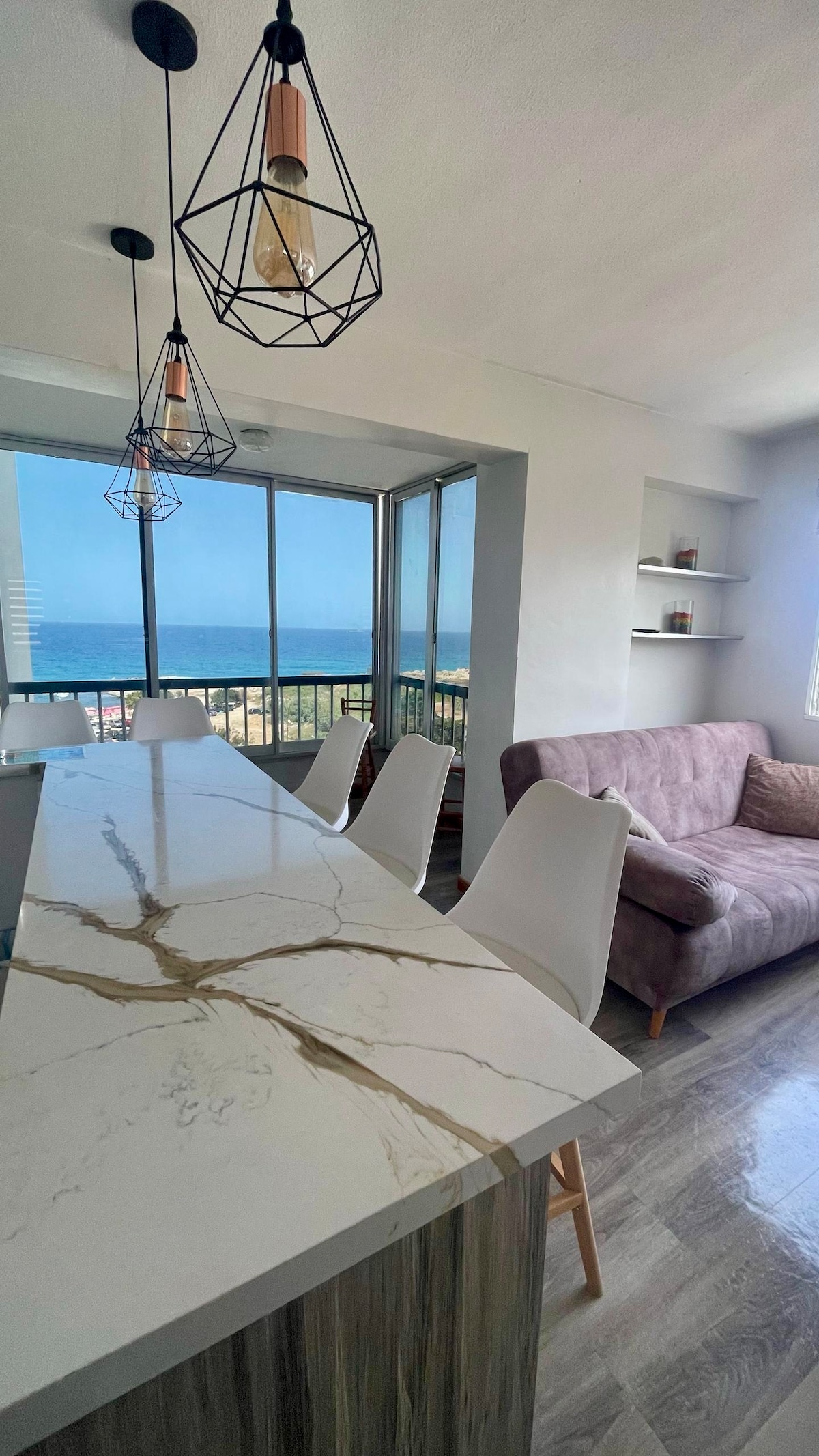 A modern bar area features a sleek countertop with a marbled finish and four white chairs. A cozy sofa in a muted tone is positioned nearby. Large windows provide a view of the ocean, filling the space with natural light.