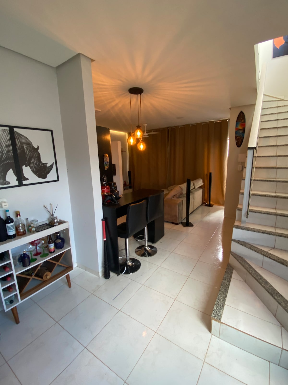 An open space is depicted, featuring a seating area with a beige sofa and a dining table. Two black bar stools align a countertop, while a decorative wine rack is positioned nearby. A staircase leads to the upper level, illuminated by a circular light fixture.