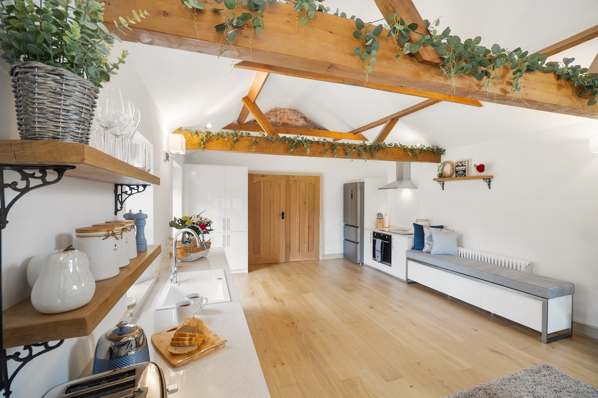 An open-plan kitchen and lounge area is highlighted by wooden beams and natural light. Shelving displays stylish kitchenware and decorative items. A cozy seating area is paired with a small table, inviting relaxation. The kitchen features modern appliances and a well-lit countertop.