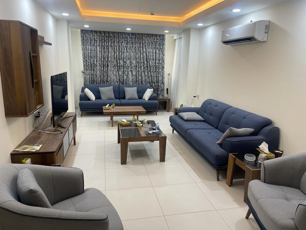 A modern living area is furnished with a mix of blue sofas and gray armchairs. A wooden coffee table occupies the center, surrounded by decorative items. Soft lighting enhances the serene atmosphere, while a large window is partially covered by patterned drapes.