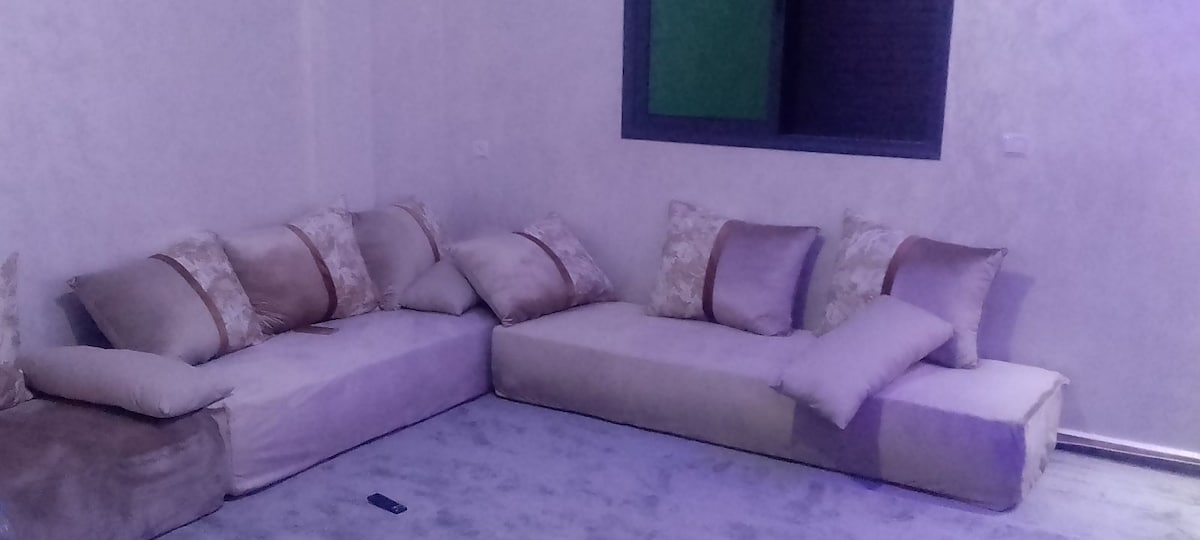 A spacious seating area features a large, L-shaped couch adorned with plush pillows. The soft upholstery is complemented by a light-colored rug beneath, creating a comfortable space for relaxation. A window with a green accent can be seen in the background.