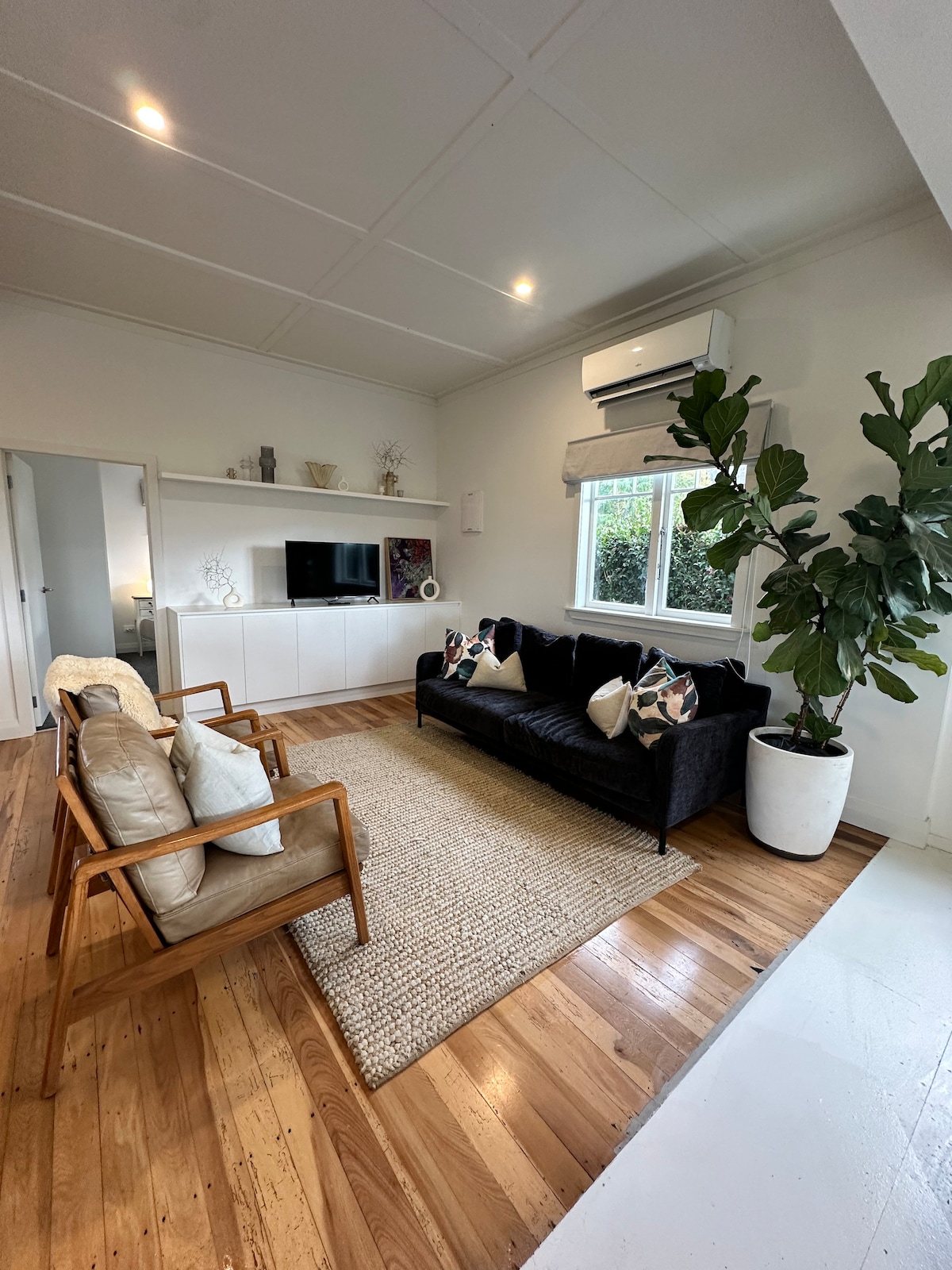 A comfortable living area features a black sofa and two wooden armchairs, complemented by decorative cushions. A large area rug is positioned on polished wooden floors. Soft lighting enhances the airy atmosphere, while a window offers views of greenery outside.