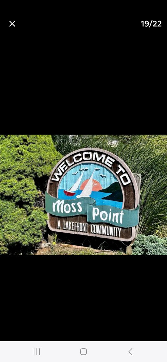 A wooden sign welcomes visitors to Moss Point, featuring a stylized image of a sailboat against a blue background. Green foliage surrounds the sign, emphasizing the natural beauty of the lakefront community.