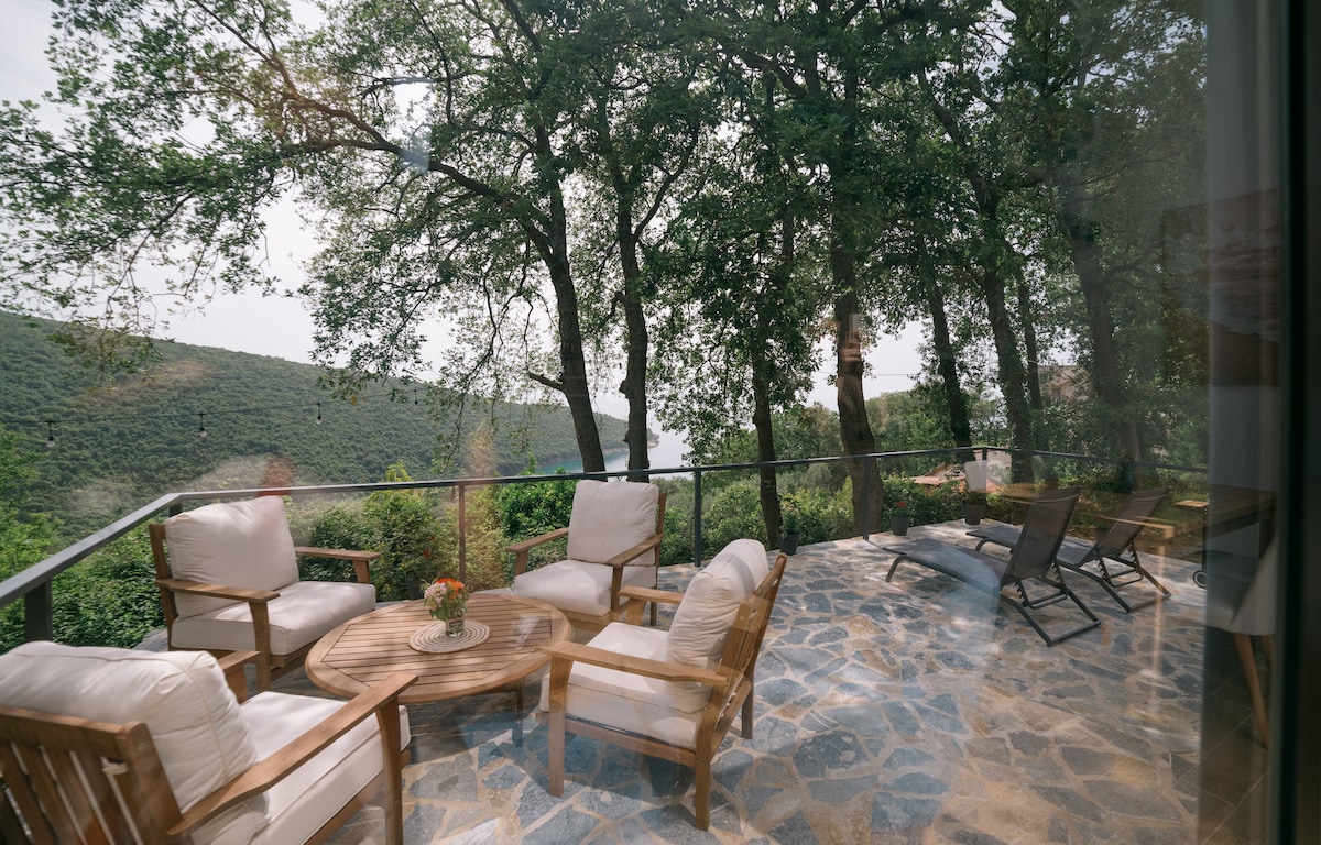 A spacious terrace features wooden furniture, including a circular table surrounded by four cushioned chairs. Two deckchairs are positioned for relaxation. Lush greenery and rolling hills are visible in the background, providing a serene setting under the shade of tall trees.