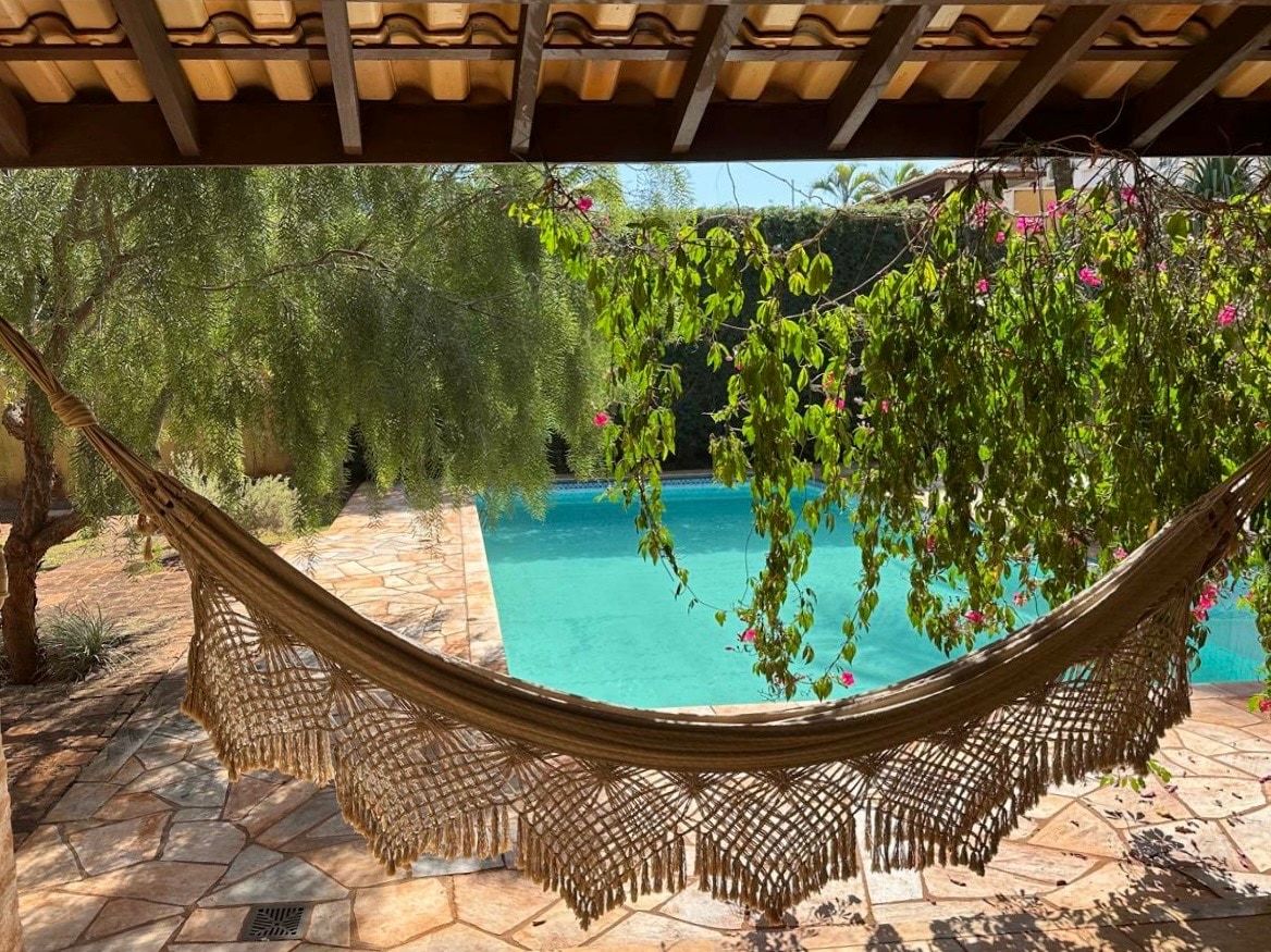 A hammock is gently draped between wooden supports, surrounded by lush greenery and vibrant flowers. The pool glimmers in the background, reflecting shades of blue under the clear sky, creating a serene outdoor setting.