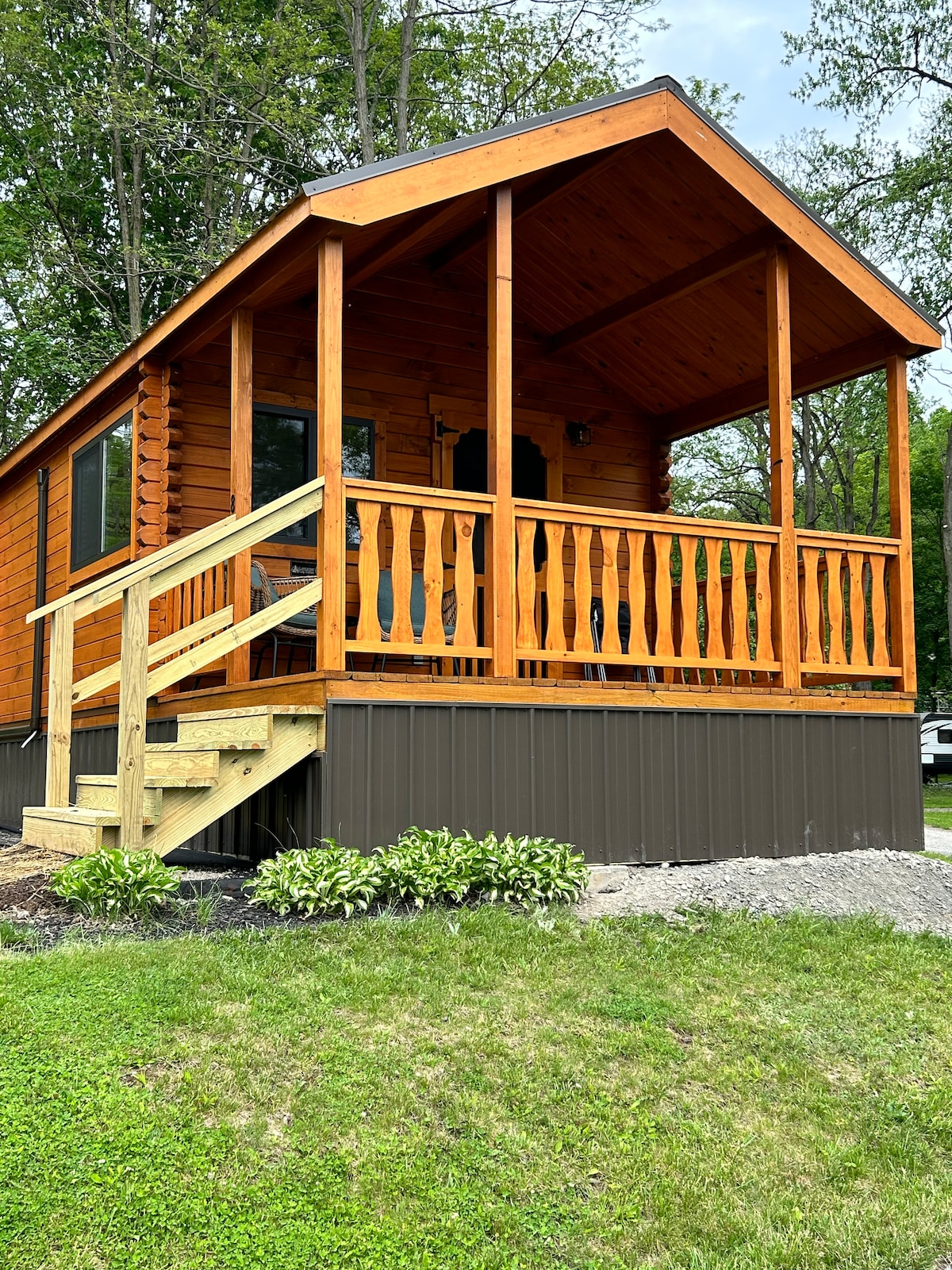 The cabin features a welcoming front porch with wooden railings and steps leading up from a landscaped area. The exterior is made of logs, complemented by large windows that offer natural light. Lush greenery surrounds the cabin, enhancing the natural setting.