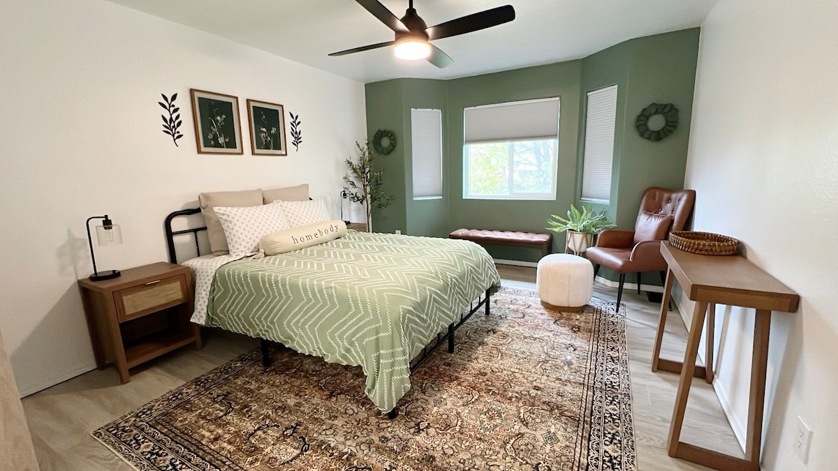 A serene bedroom is highlighted by a spacious bed adorned with a soft green coverlet. Natural light filters through dual windows framed by white blinds. A modern chair and small bench provide additional seating, while decorative plants and wall art enhance the inviting ambiance.