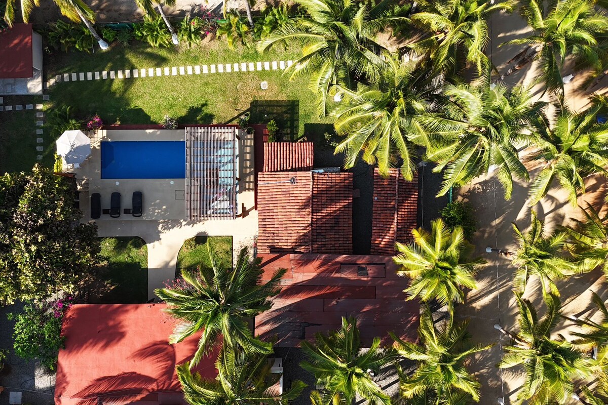 An aerial view captures a vibrant landscape, featuring a clear blue pool surrounded by lush greenery and palm trees. A charming red-roofed accommodation is nestled among expansive gardens, with a pathway bordered by tropical plants leading to open spaces.