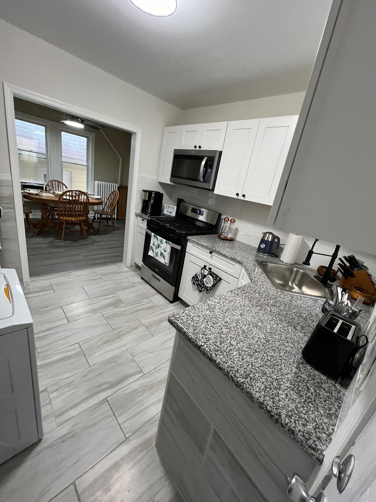 The kitchen is equipped with modern appliances, including a microwave and an oven. A granite countertop enhances the workspace, while white cabinetry offers storage. An open doorway leads to a dining area, featuring wooden chairs and a table visible through a window.