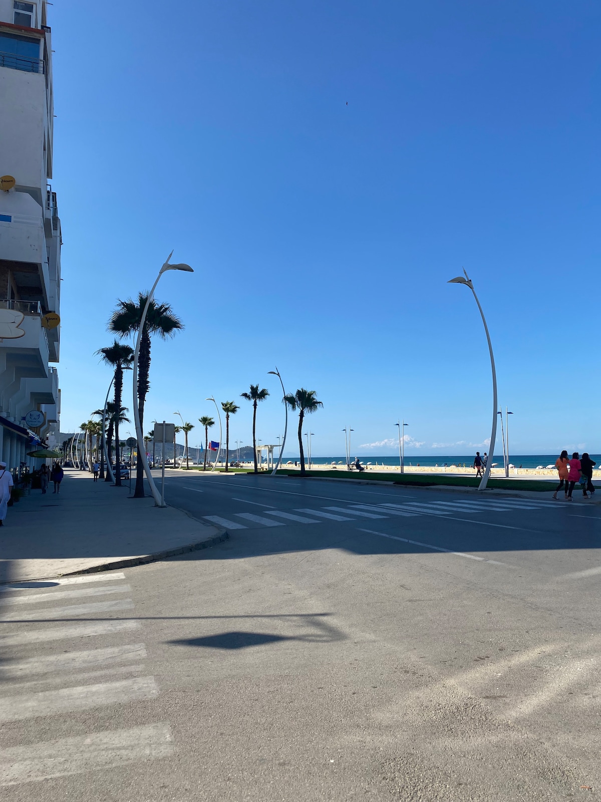 A wide street lined with palm trees leads towards the beach, featuring modern street lamps. The expansive blue sky is visible above, with hints of the ocean peeking in the distance. Sidewalks are present, allowing for pedestrian access along the vibrant promenade.