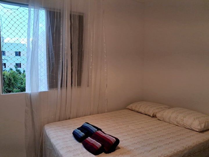 Spavaća soba s bračnim krevetom, stropnim ventilatorom, prijenosnim ventilatorom, ormarom, radnom površinom, ogledalom iza vrata i zavjesom na prozoru (sa zaštitnom mrežicom).