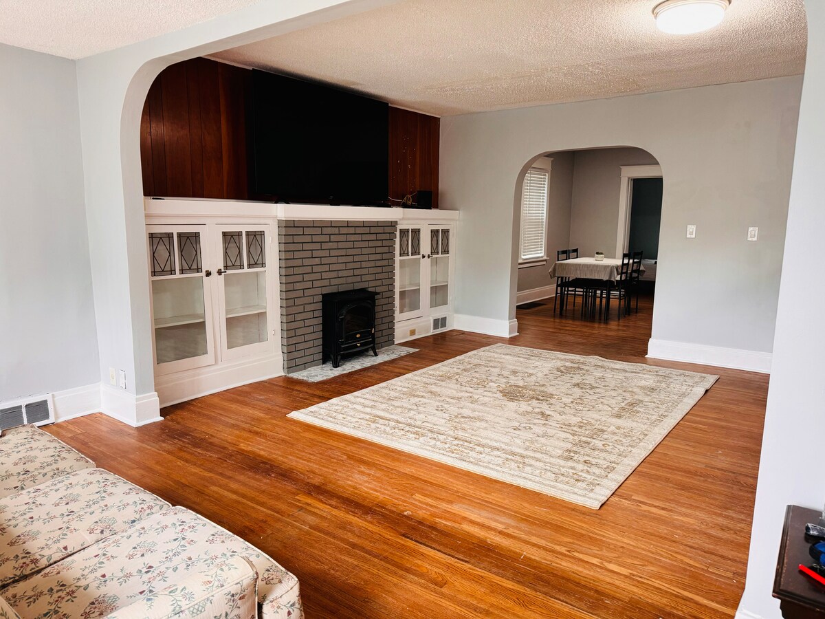 An open-concept living area features rich hardwood flooring. A neutral area rug lies centered on the floor, while a sleek fireplace and white cabinetry provide a focal point. The wall opposite houses a large television, with a dining area visible through a doorway.