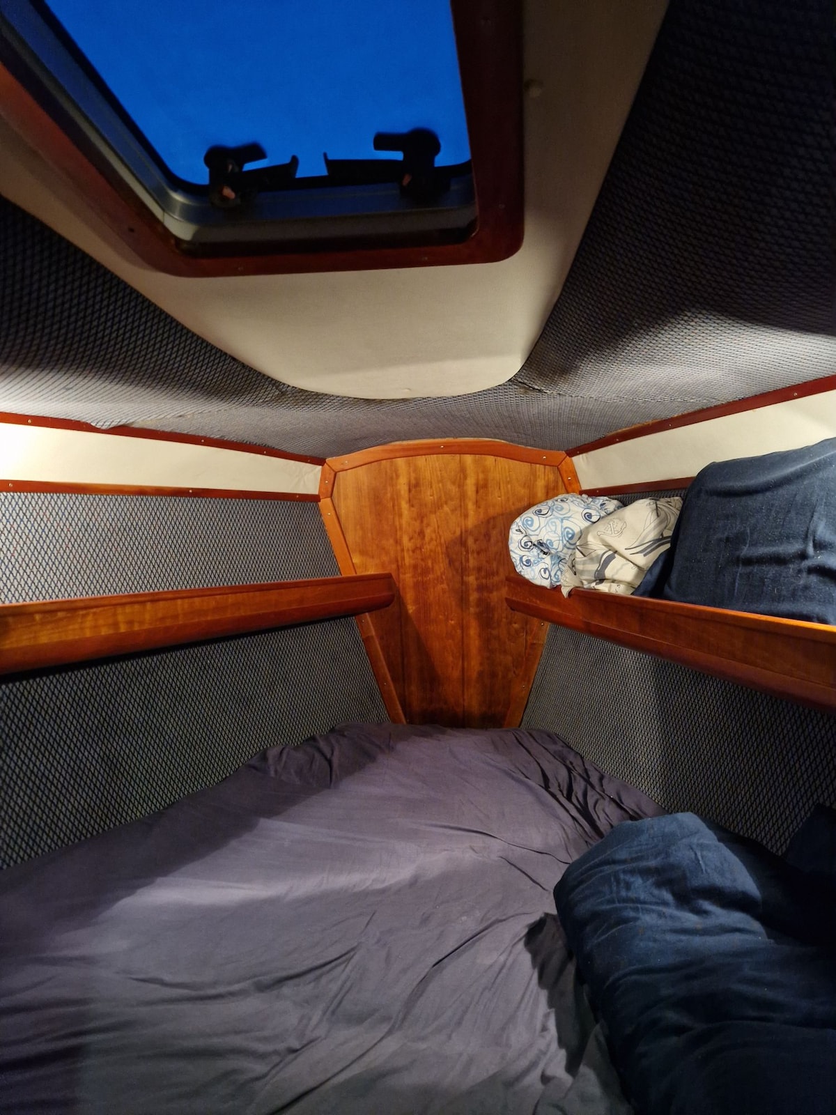 A cozy sleeping area within the boat features a spacious bed dressed in soft bedding. The wooden paneling provides a warm contrast to the fabric details. A small window above allows natural light, illuminating the finely woven textures of the walls.
