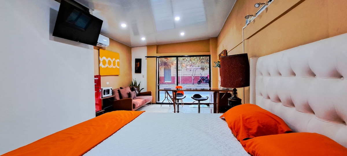 A studio apartment features a spacious bed with orange accent pillows. A comfortable seating area is arranged near a large window, allowing natural light to brighten the space. A small dining table is visible, complemented by modern lighting and decor.