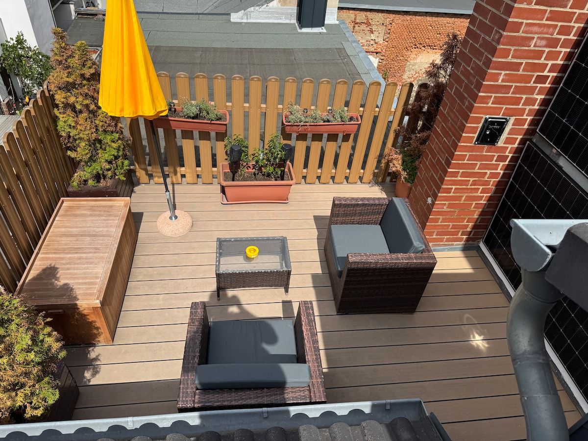 A private terrace is shown, featuring a wooden deck with outdoor seating arrangements in a modern style. Potted plants add greenery, while an umbrella provides shade. The space is well-suited for relaxation, with a small table positioned at the center.