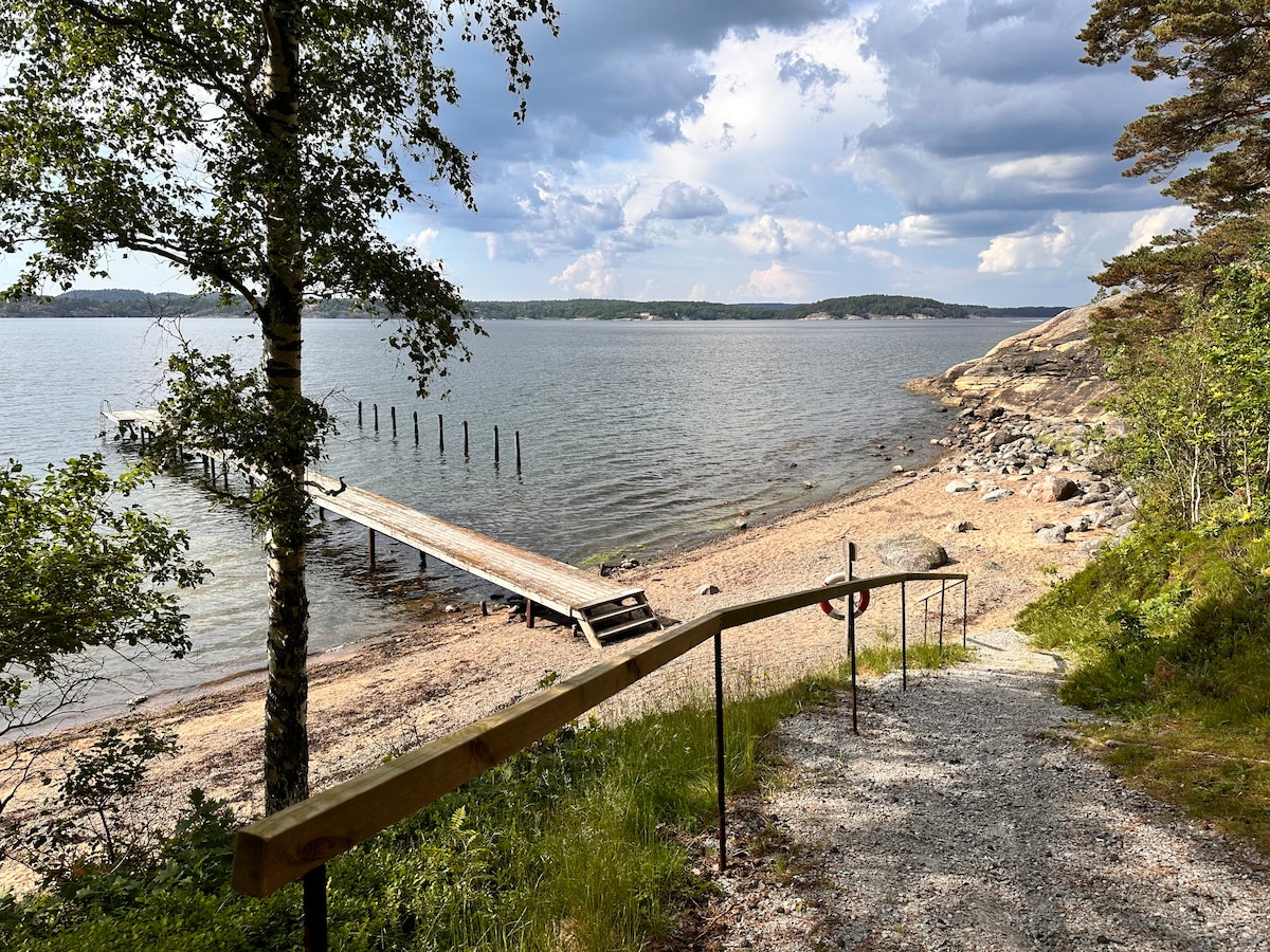 A sandy beach is framed by lush greenery and rocky shores, with a wooden pier extending into calm waters. A gentle slope is marked by a handrail leading down from a gravel path, inviting access to the scenic waterfront.