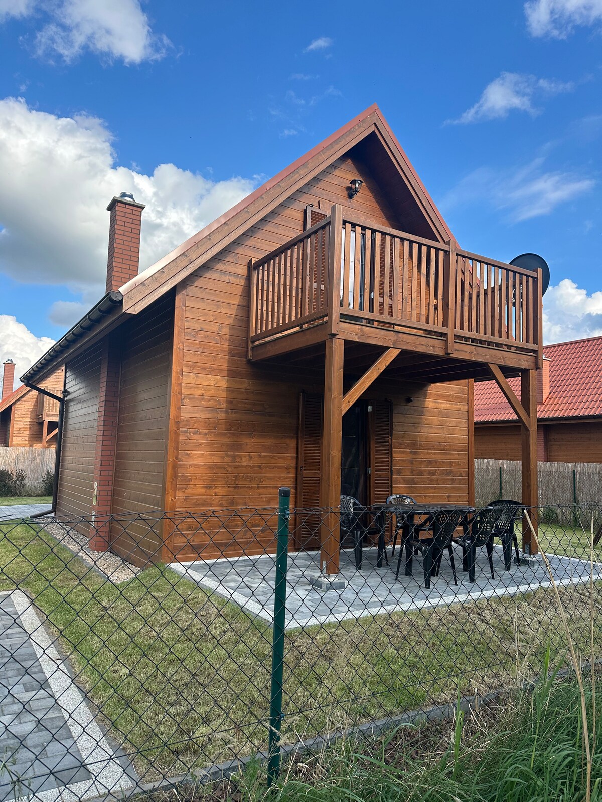 A wooden cabin is shown with a balcony accessed by a set of stairs. Black outdoor chairs surround a table on a stone patio. Lush green grass borders the cabin, and a fence encloses the property, while nearby structures and a blue sky with clouds can be seen.