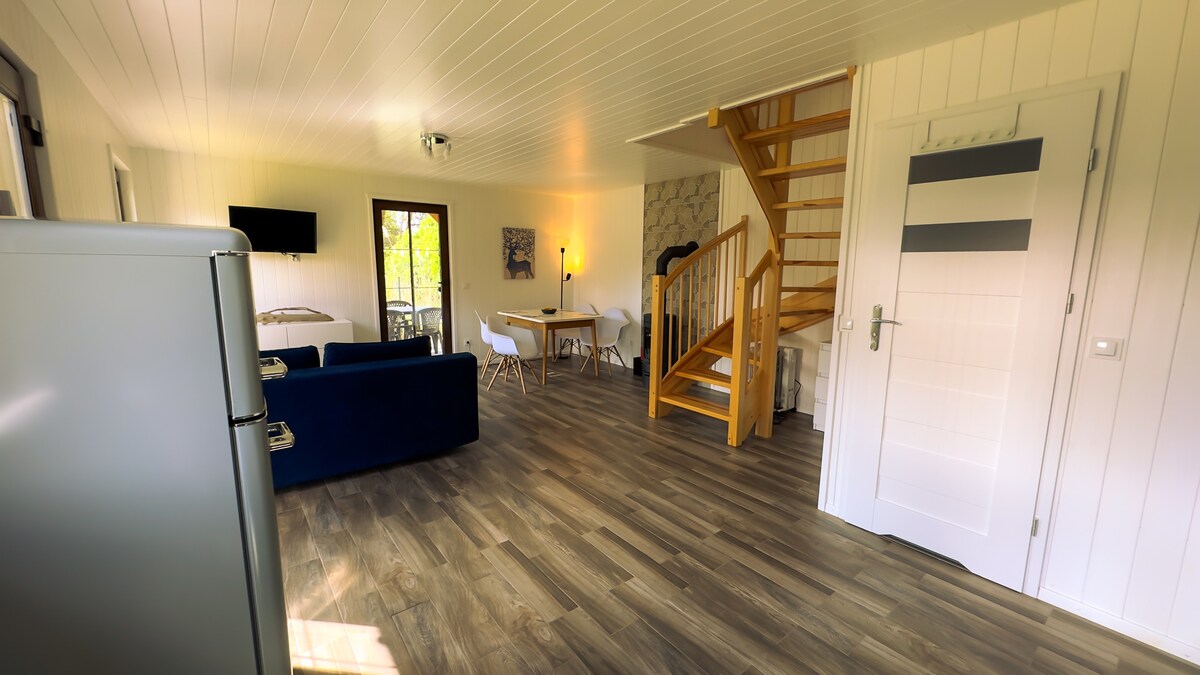 An open living space is showcased, featuring a modern kitchen, a dining area with a round table, and a cozy seating arrangement. Natural light enters through large windows, illuminating the wood-patterned flooring. A staircase with wooden steps is visible, leading to an upper level.