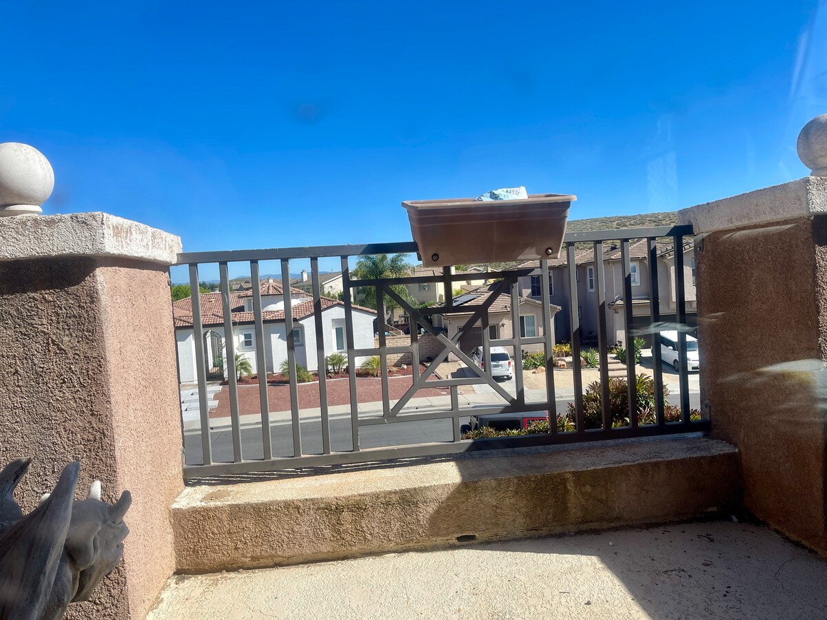 A view from an attached balcony shows a metal railing and a planter box. The surrounding neighborhood features residential buildings with tile roofs and landscaped areas. A clear blue sky provides a bright backdrop, enhancing the serene setting.