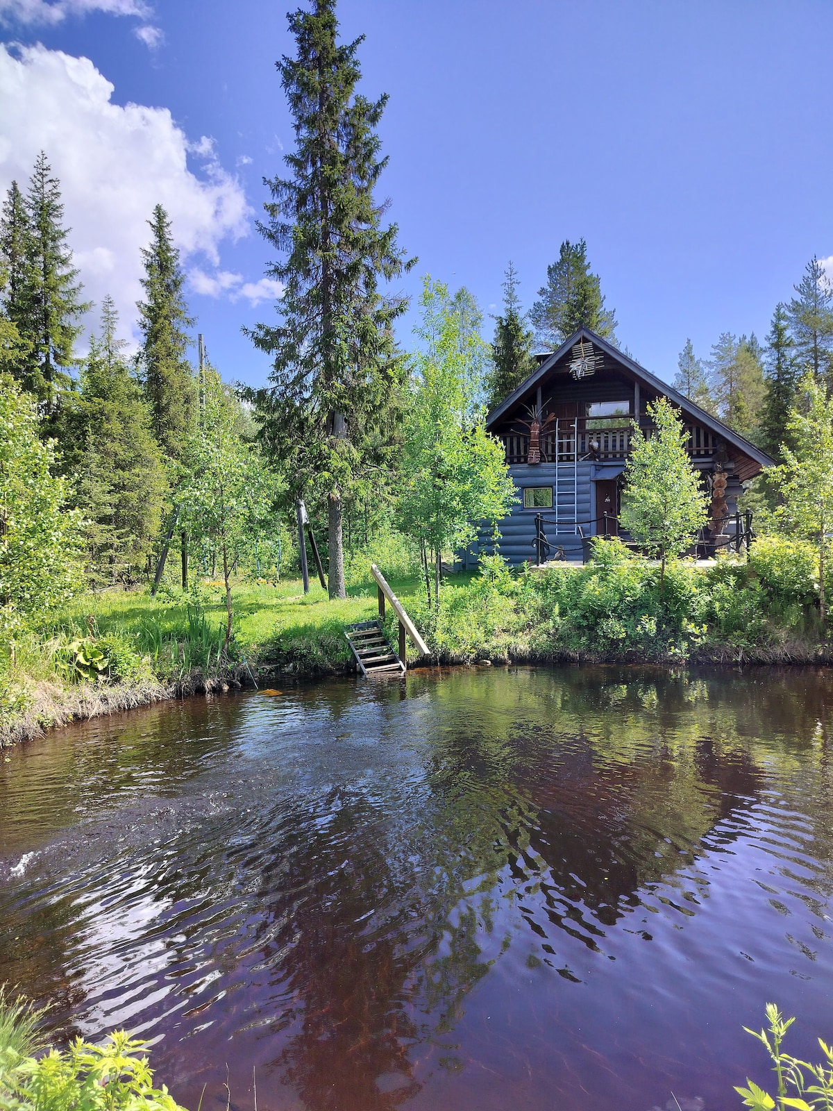 A wooden cabin is nestled among trees, with greenery surrounding it. A staircase leads to the water's edge, where a calm body of dark water reflects the blue sky and clouds. The structure features a sloped roof and a covered balcony.