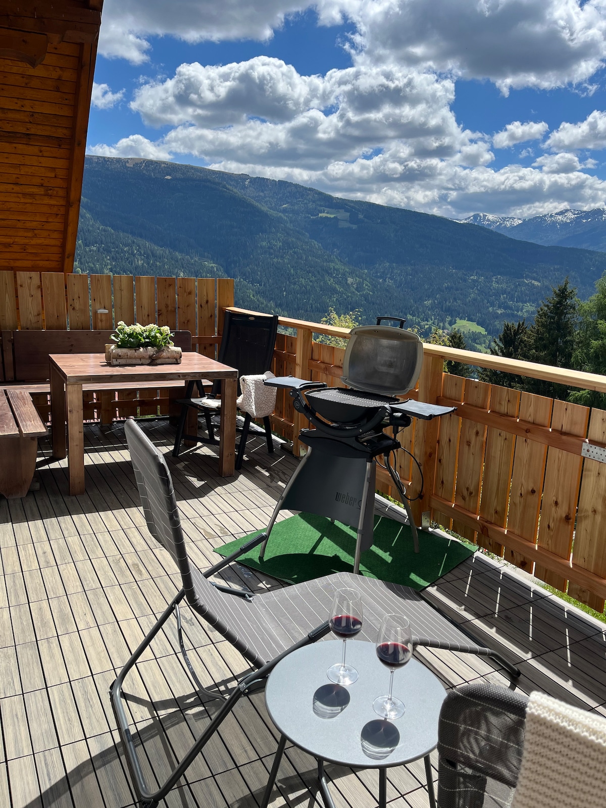 An outdoor terrace is presented with a wooden table and chairs, accompanied by a grill. Two glasses of red wine sit on a small round table, highlighting the setting. The backdrop features expansive views of mountains and valleys under a partly cloudy sky.