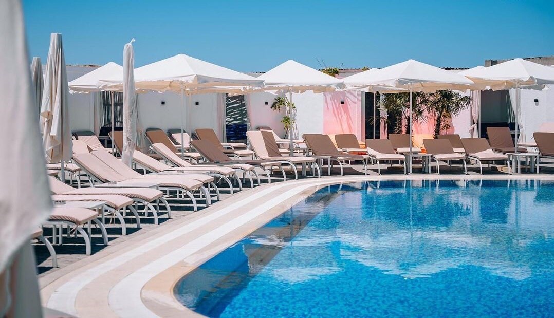 A large resident-only pool is surrounded by a sun terrace equipped with numerous lounge chairs. White umbrellas provide shade for the sunbathers. The clear blue water reflects the bright sky, enhancing the serene atmosphere of the outdoor leisure space.
