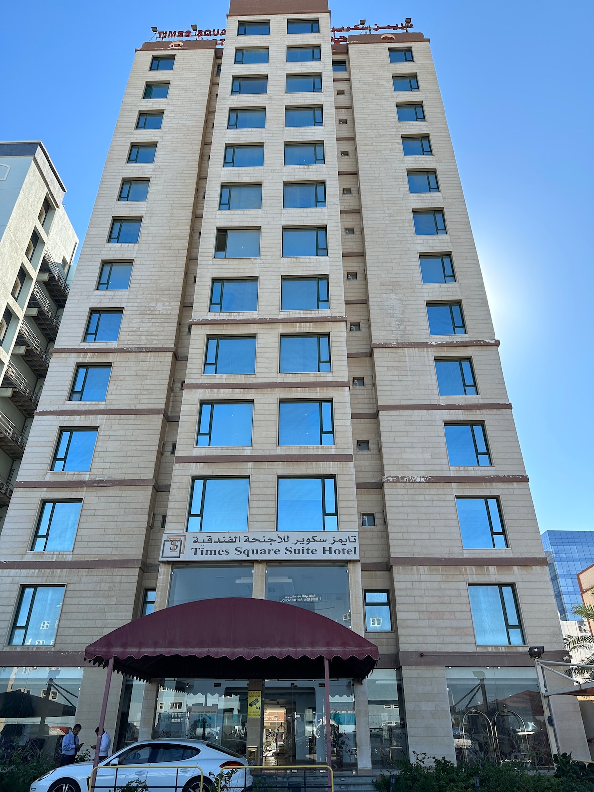 A modern ten-story building features large glass windows that reflect the clear blue sky. The entrance is marked by a covered awning, and a sign indicates the name of the establishment. A parked car is visible in front, adding to the urban ambiance.