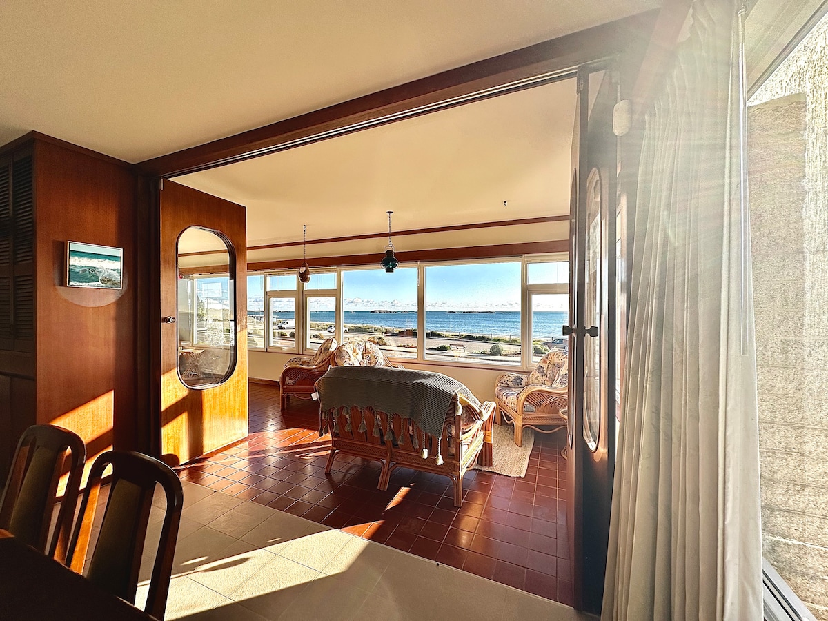 An inviting indoor space features large windows that provide a clear view of the ocean. A wooden seating area with light-colored upholstery is positioned to enjoy the scenery. Natural light enhances the warm tones of the room, creating a bright atmosphere.