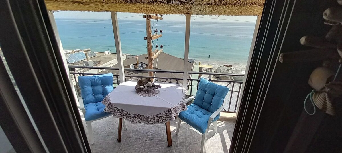 A bright outdoor seating area features a round table with a white tablecloth. Two blue cushioned chairs are positioned around the table. The backdrop showcases a clear view of the ocean, with gentle waves visible and the horizon line stretching into the distance.
