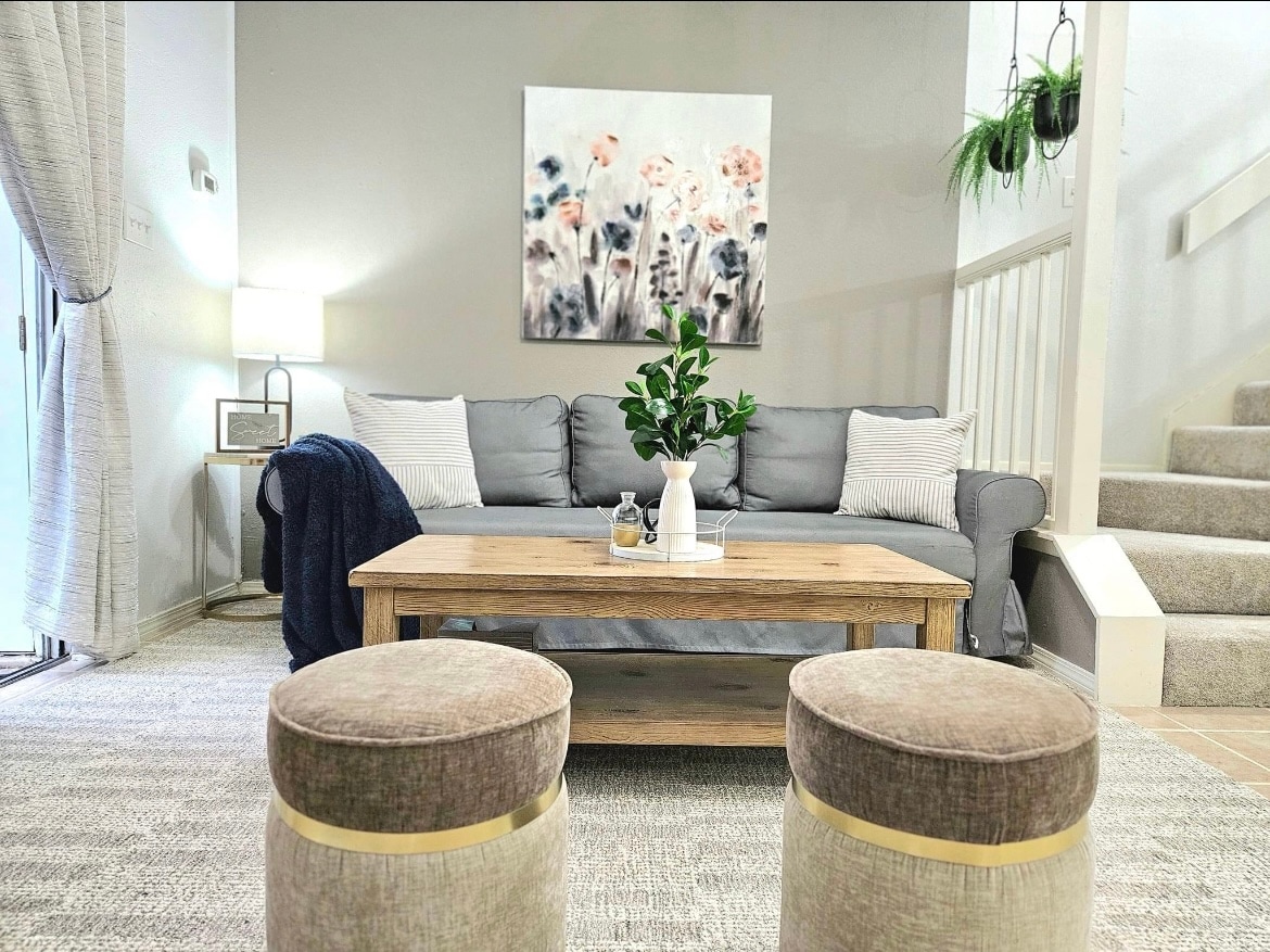 A living area features a light grey sofa adorned with decorative cushions, set against a neutral wall. A coffee table is positioned centrally, decorated with a plant and a white vase. Two upholstered stools are placed nearby, with natural light filtered through sheer curtains.
