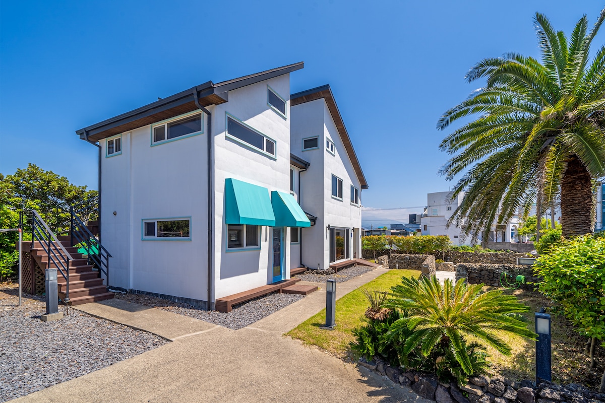 A modern two-story building is set amidst lush greenery, featuring a well-maintained yard. Large windows and an awning provide natural light and shade. A staircase leads to the entrance, while surrounding palm trees enhance the tranquil atmosphere.