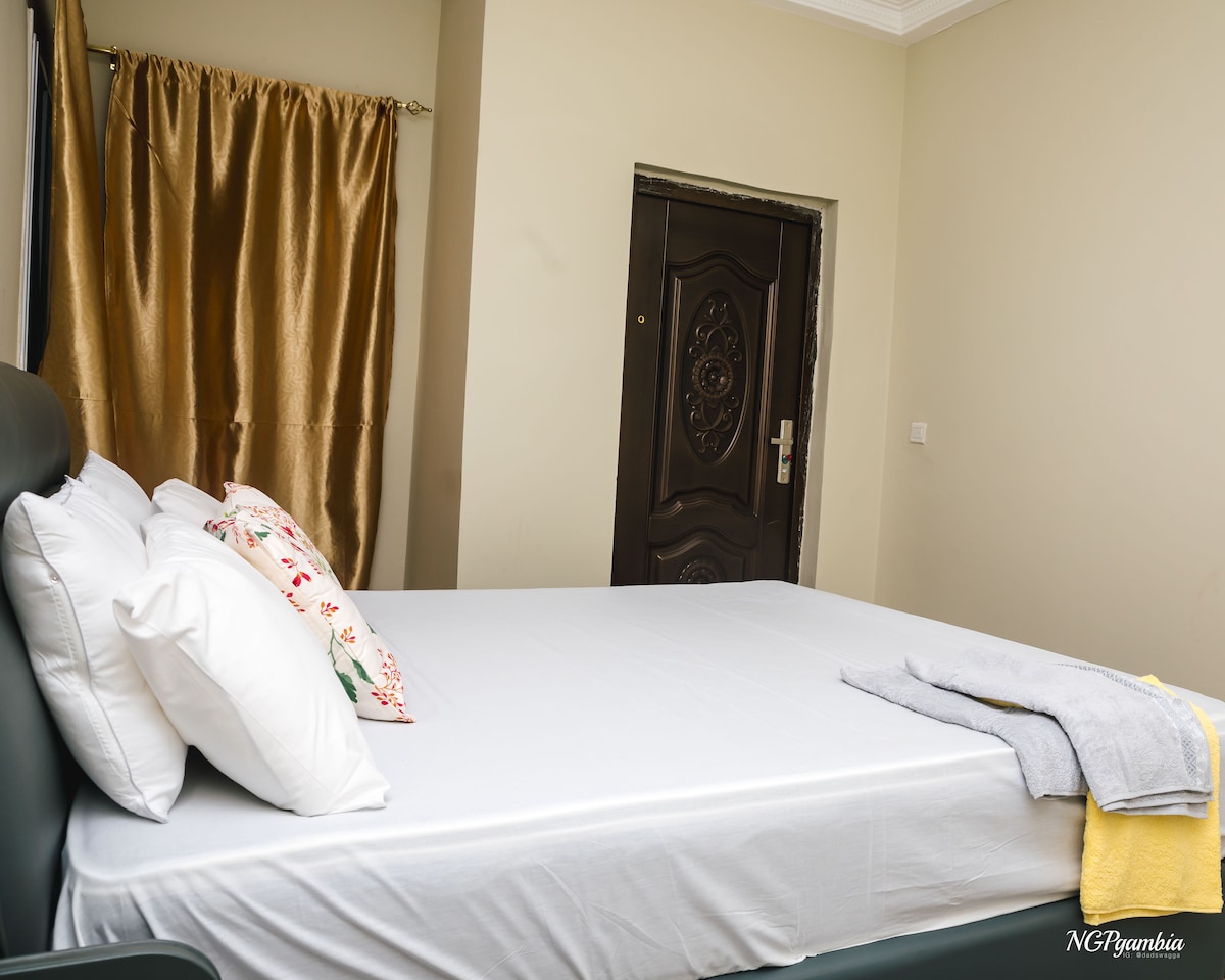 A spacious bed is positioned against a neutral wall, dressed in crisp white bedding. Decorative pillows are arranged neatly at the head of the bed. Soft, golden curtains frame a window, and a wooden door with an intricate design is visible in the background.