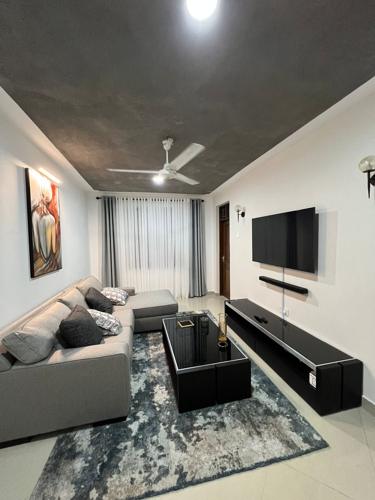A modern living area is showcased, featuring a comfortable gray sectional sofa adorned with dark accent pillows. A sleek black coffee table is positioned in front, with a decorative item placed atop. Natural light filters through sheer curtains, adding to the inviting ambiance.