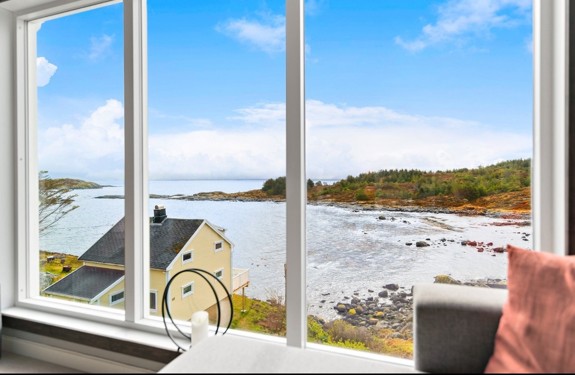 A waterfront view is showcased through large glass windows, revealing a serene landscape of calm waters and rocky shoreline. Distant trees and a yellow house are visible against the backdrop of a partly cloudy sky.