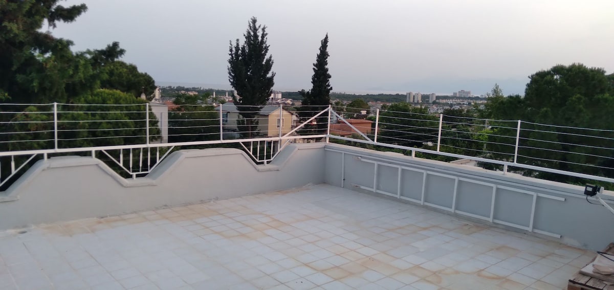 A spacious rooftop terrace features a clear view of the surrounding landscape, including distant trees and buildings. The area is enclosed with a white railing, providing safety and an open feel. The tiled surface is unadorned, allowing for various outdoor activities.