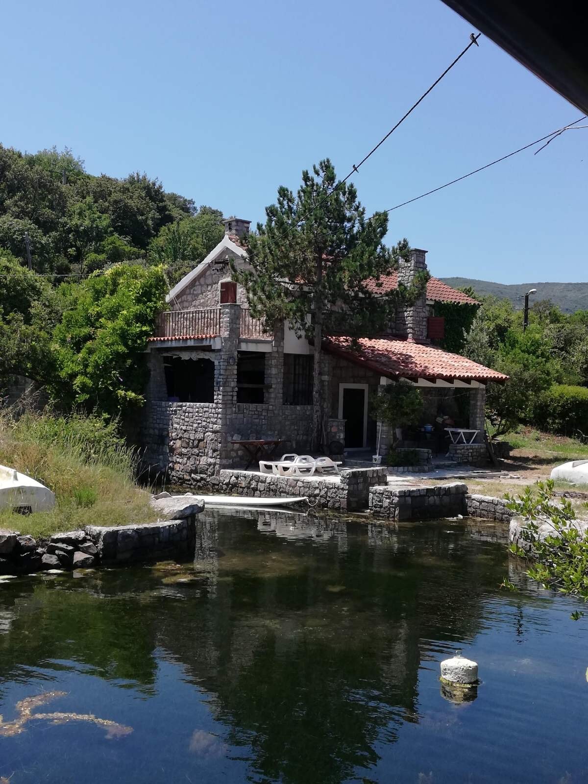 A rustic stone house is set amidst lush greenery, featuring a red-tiled roof and multiple windows. A clear pond reflects the structure and surrounding landscape, while a small seating area with white tables and chairs is visible nearby.