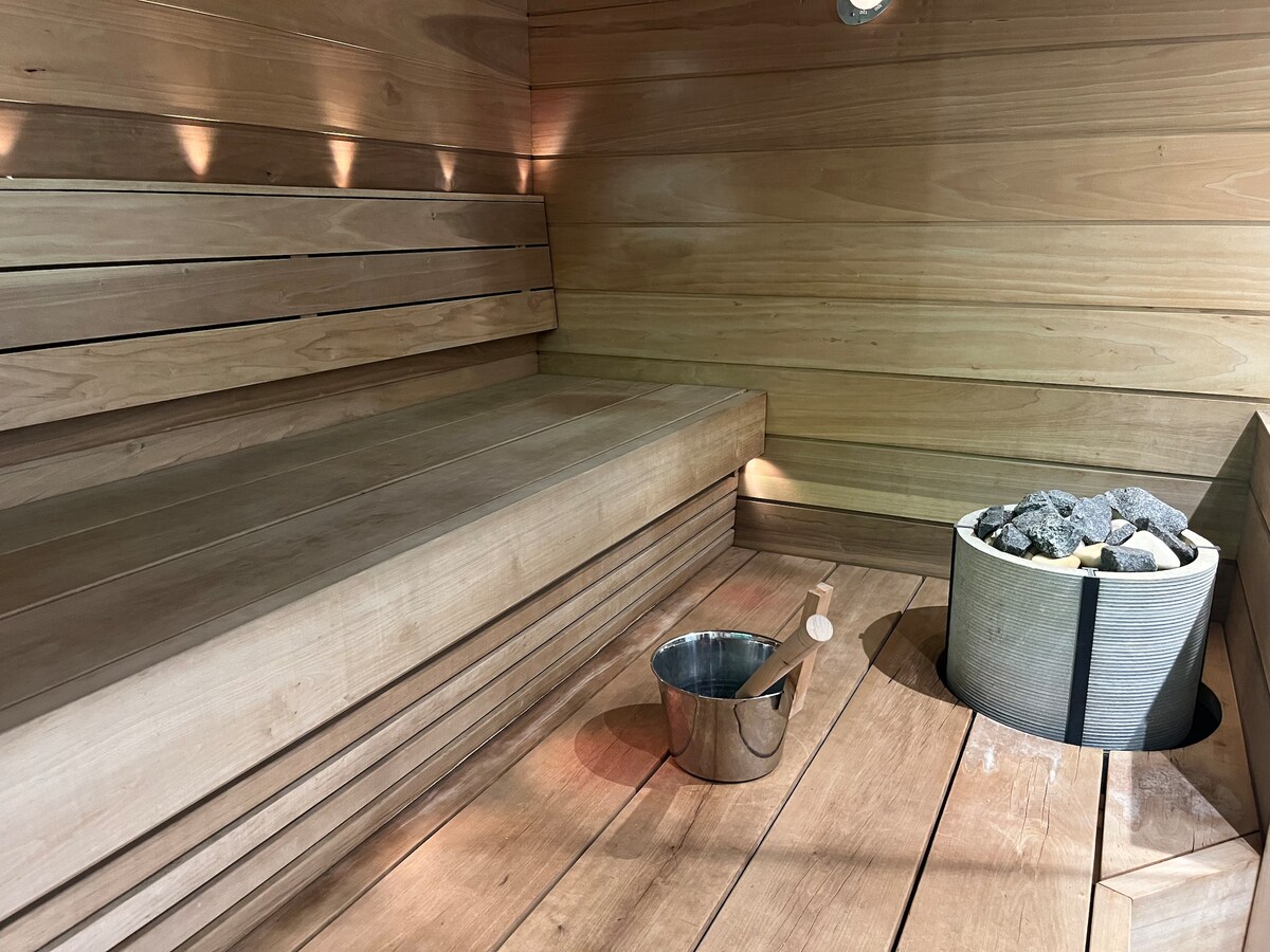 A wooden sauna interior features smooth cedar benches, providing ample seating. A stone-filled heater is positioned centrally, surrounded by a circular stone base. A metal bucket rests on the floor, offering convenience for water use during the sauna experience.